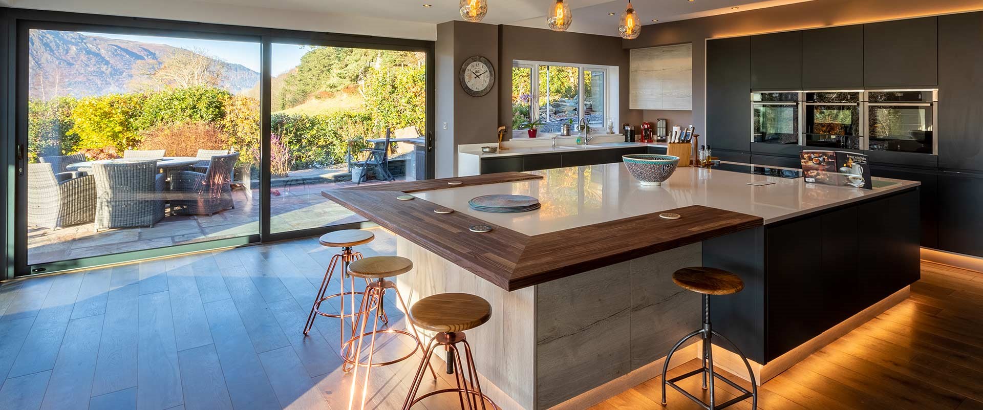 A matte black kitchen with a dark wood breakfast bar with lowlights and a view over hills.